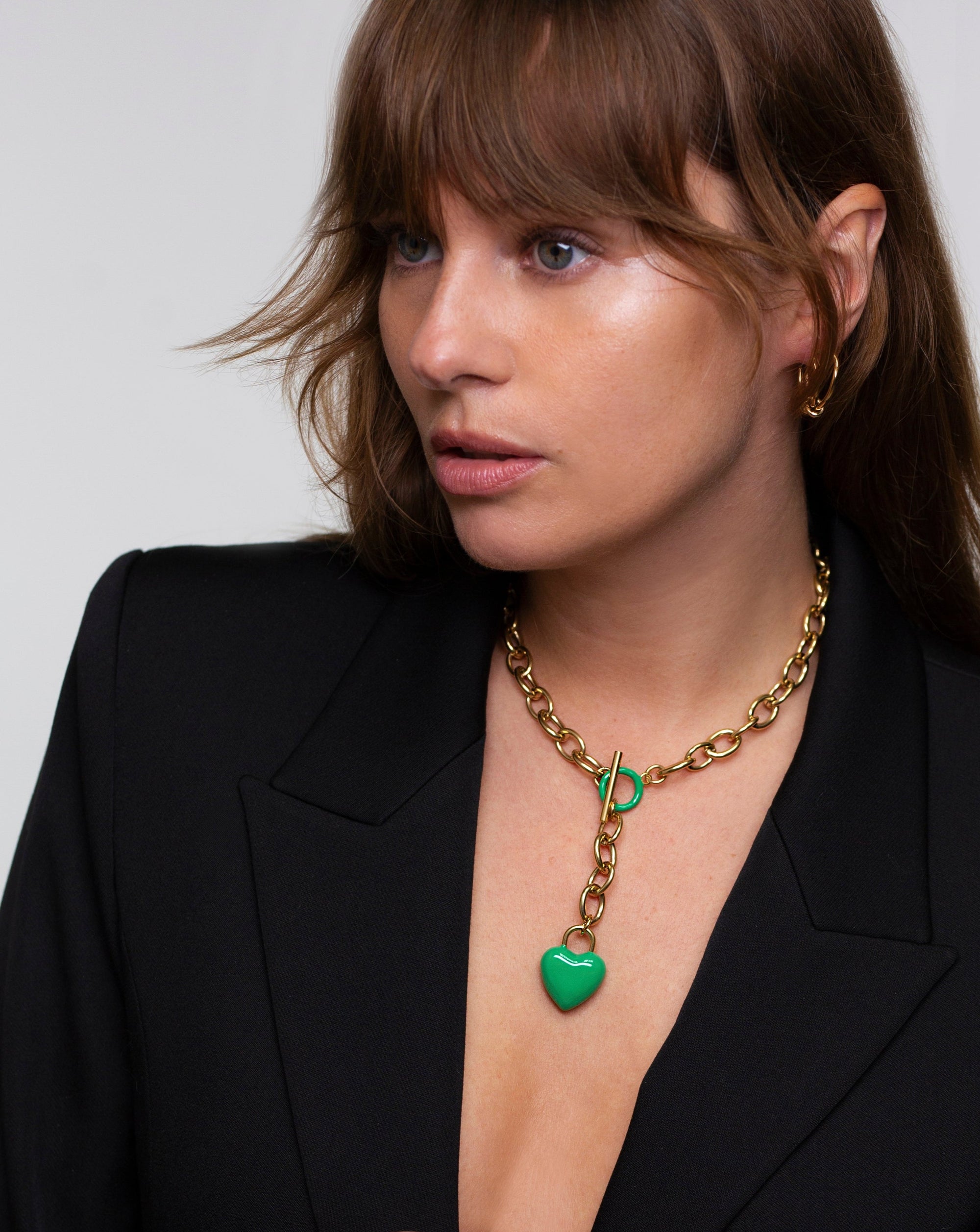 A person with brown hair and a neutral expression wears a black blazer and a chunky gold necklace with an enamel-coated heart-shaped pendant from For Art's Sake®, called The Kiss Necklace Pink. The individual also has a gold hoop earring visible on the right ear. The background is plain white.