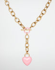 The Kiss Necklace Pink by For Art's Sake® features a pink, enamel-coated heart-shaped pendant hanging from a central T-bar clasp, which is also adorned with a pink ring.