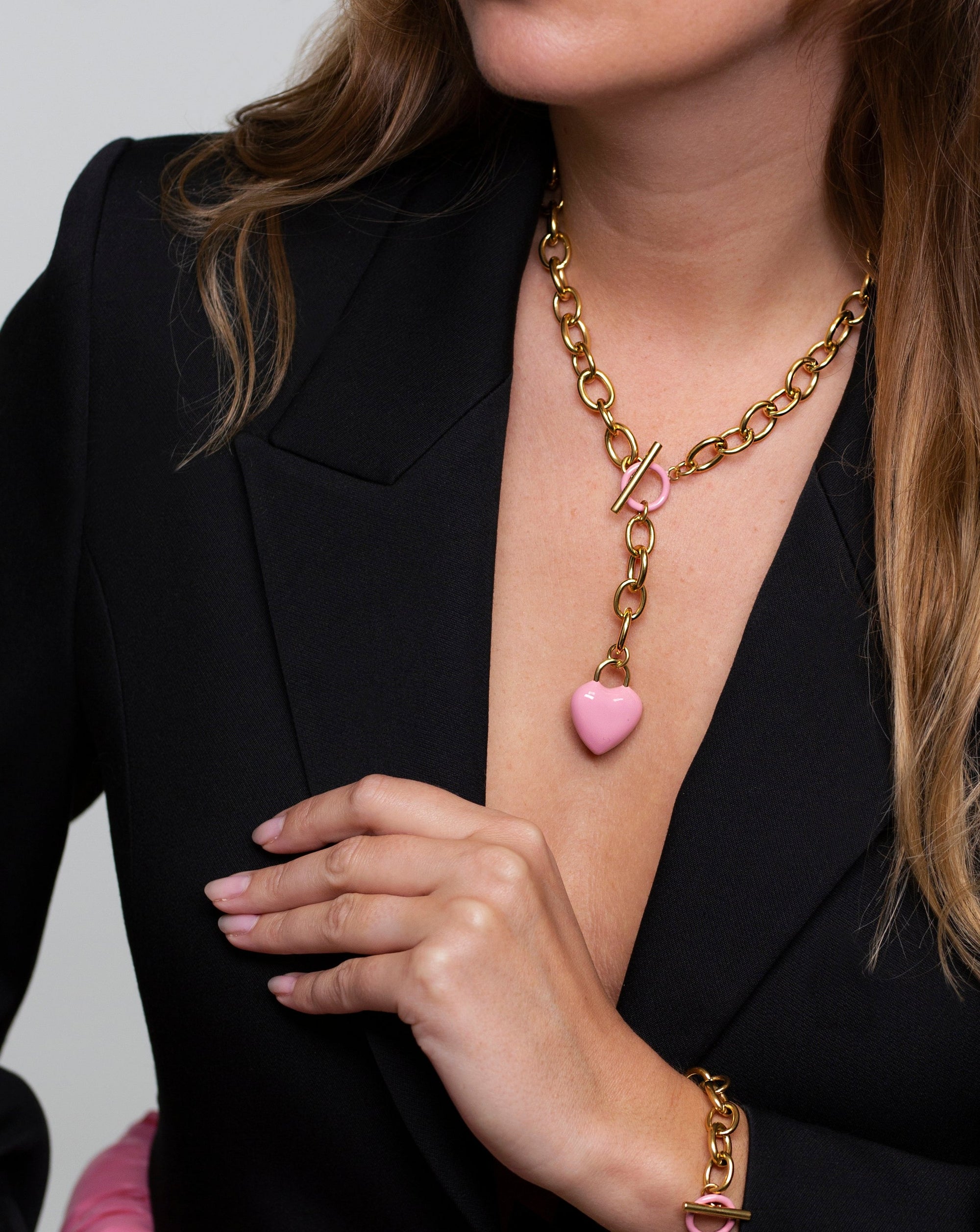 A woman wearing a black blazer is seen with The Kiss Necklace Pink from For Art's Sake®, featuring an enamel-coated heart pendant and a T-bar closure. She is also adorned with a matching gold bracelet with pink details, her hand resting gracefully on her chest.