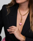 A woman wearing a black blazer is seen with The Kiss Necklace Pink from For Art's Sake®, featuring an enamel-coated heart pendant and a T-bar closure. She is also adorned with a matching gold bracelet with pink details, her hand resting gracefully on her chest.