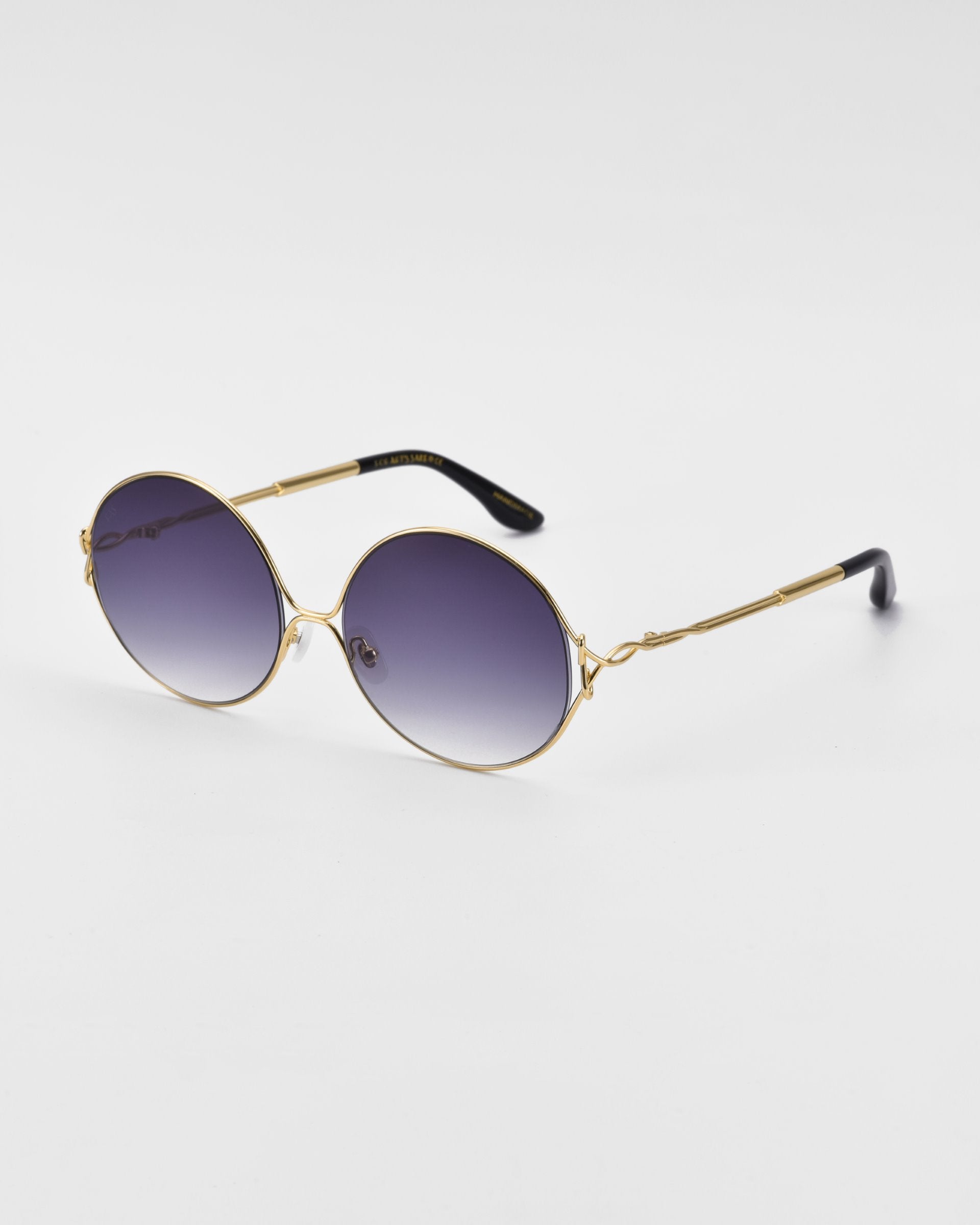 A pair of sleek, retro-inspired For Art's Sake® Aura sunglasses with gold-colored metal frames and gradient dark lenses are placed on a plain white background. The temples are thin with black tips, and the overall design is minimalist and stylish.