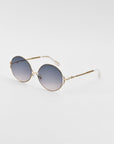 A pair of retro-inspired Aura sunglasses by For Art's Sake® with oversized round frames in gold metal and gradient blue lenses. The Aura sunglasses have thin temples with white tips and a minimal design against a plain white background.