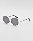 A pair of For Art's Sake® Aura retro-inspired sunglasses with gradient tinted lenses and a thin, silver metal frame lies on a white surface. The temples are black at the ends.