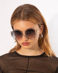 A young person with medium-length, light brown hair is wearing large, stylish For Art's Sake® Sapphire sunglasses with UV protection and hoop earrings. They have a serious expression and are dressed in a sheer black top. The background is plain and white.