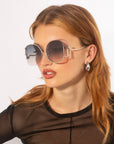 A person with long, light brown hair is wearing large, round For Art's Sake® Sapphire sunglasses with gradient lenses and silver frames offering UV protection. They are also wearing a sheer black top and silver hoop earrings. The background is plain white.