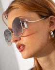 Woman wearing large round For Art's Sake® Sapphire sunglasses with silver frames and rectangular decorative accents gazes off to the side. She has light brown hair and wears large silver hoop earrings. The lenses, featuring an anti-reflective coating, offer superior UV protection. The background is a blurred, neutral shade.