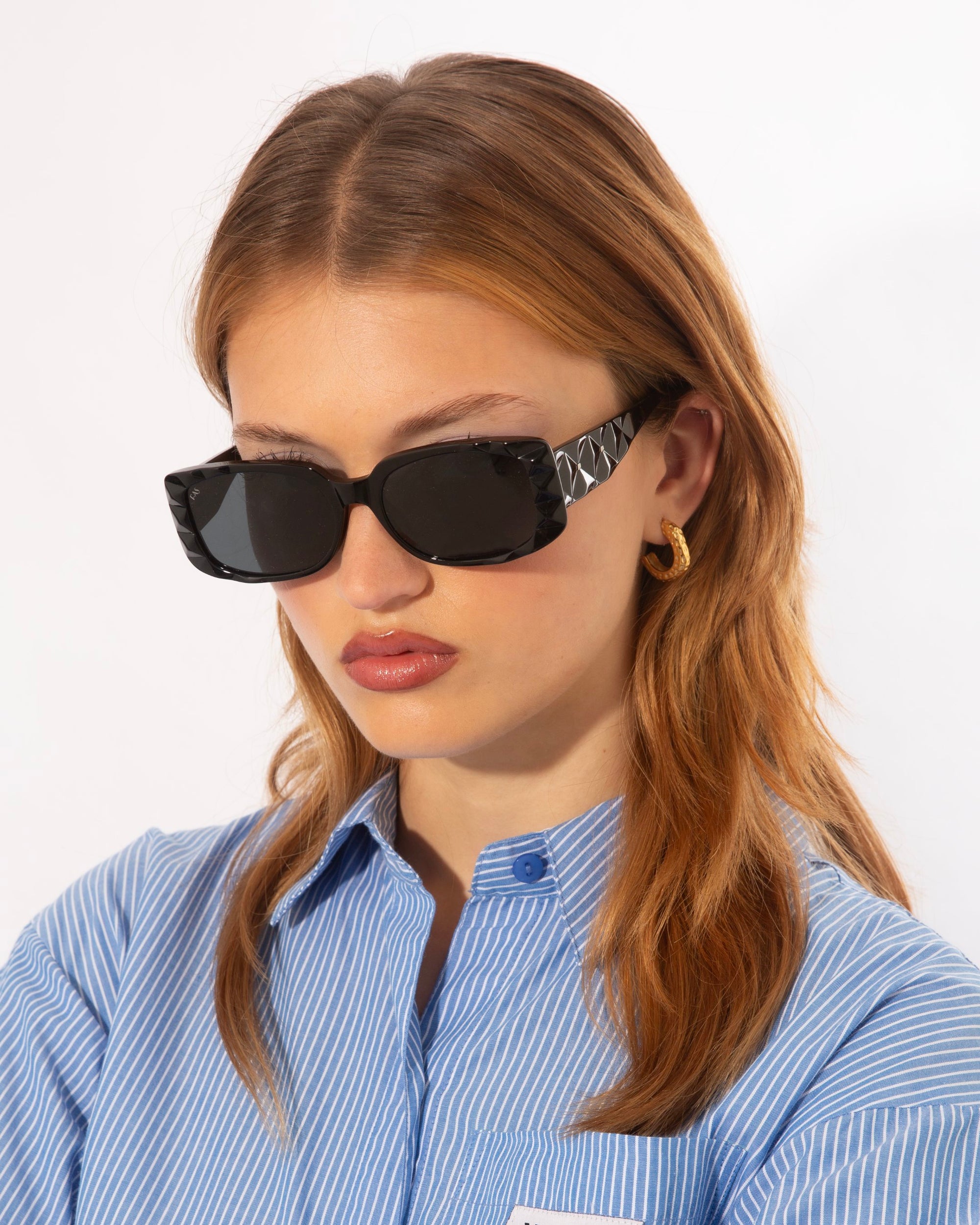 A person with wavy, auburn hair is wearing black rectangular For Art's Sake® Cushion sunglasses with a textured frame and ultra-lightweight nylon lenses offering 100% UV protection. They also have gold hoop earrings and a blue, striped button-up shirt. The person is looking down with a serious expression against a plain white background.