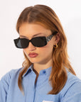 A person with wavy, auburn hair is wearing black rectangular For Art's Sake® Cushion sunglasses with a textured frame and ultra-lightweight nylon lenses offering 100% UV protection. They also have gold hoop earrings and a blue, striped button-up shirt. The person is looking down with a serious expression against a plain white background.