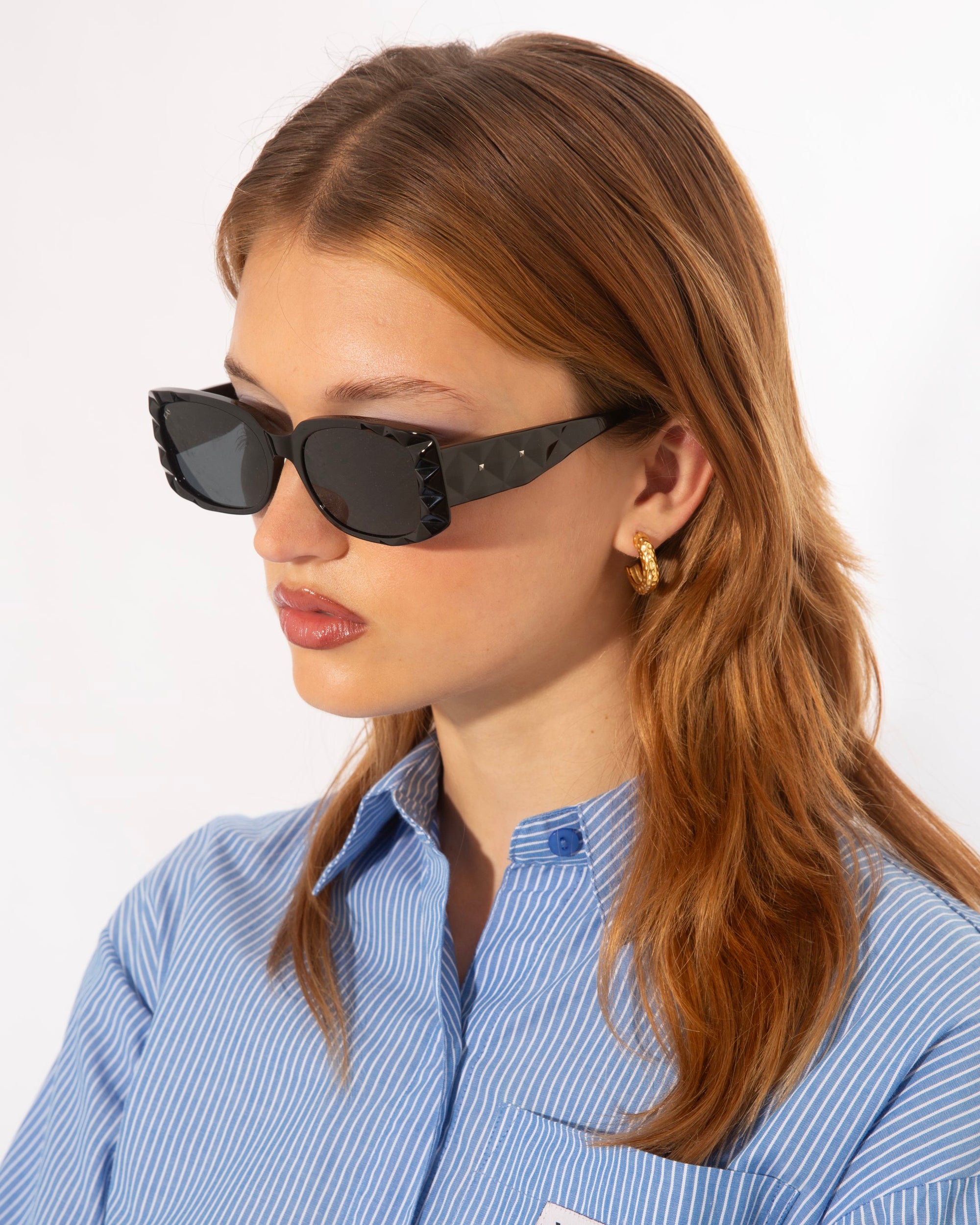 A person with light brown hair wearing black rectangular For Art's Sake® Cushion sunglasses with Ultra-lightweight Nylon lenses, a blue and white striped button-up shirt, and gold hoop earrings stands in front of a plain white background.