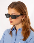 A person with light brown hair wearing black rectangular For Art's Sake® Cushion sunglasses with Ultra-lightweight Nylon lenses, a blue and white striped button-up shirt, and gold hoop earrings stands in front of a plain white background.