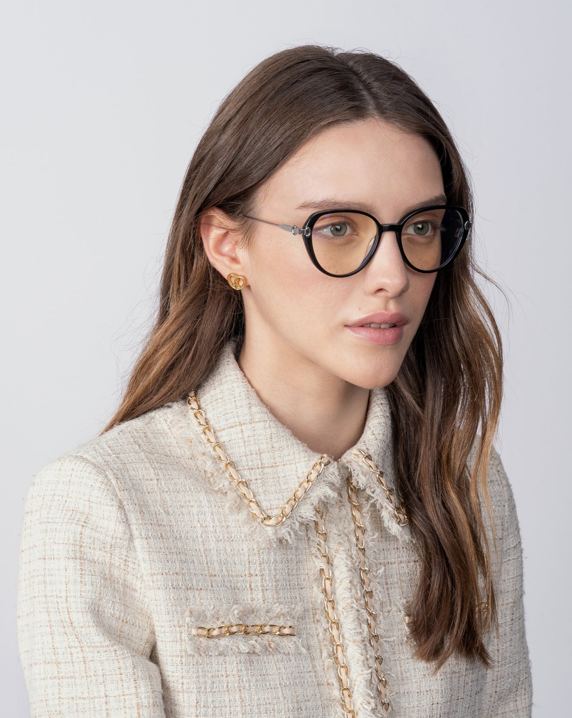 A woman with long, brown hair wearing For Art's Sake® Waterhouse glasses with prescription lenses and a beige textured jacket adorned with 18-karat gold-plated chain detailing. She has small gold stud earrings and is looking slightly to the side against a plain, light gray background.