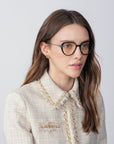 A woman with long, brown hair wearing For Art's Sake® Waterhouse glasses with prescription lenses and a beige textured jacket adorned with 18-karat gold-plated chain detailing. She has small gold stud earrings and is looking slightly to the side against a plain, light gray background.