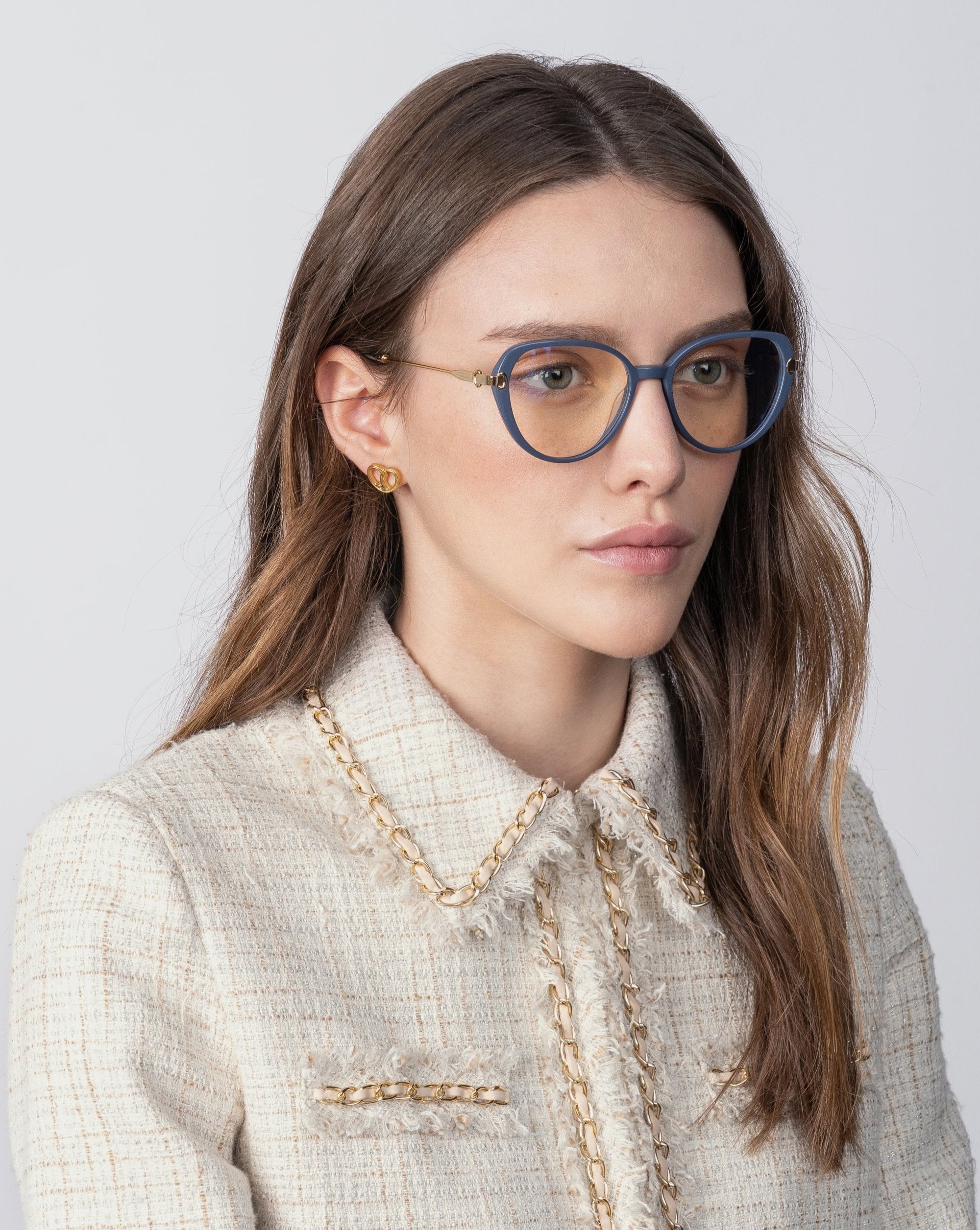 A person with light skin and long, brown hair is wearing round blue Waterhouse glasses with prescription lenses and gold earrings by For Art's Sake®. They have on a textured, off-white blazer with 18-karat gold-plated chain details and a high collar. The background is plain and light in color.