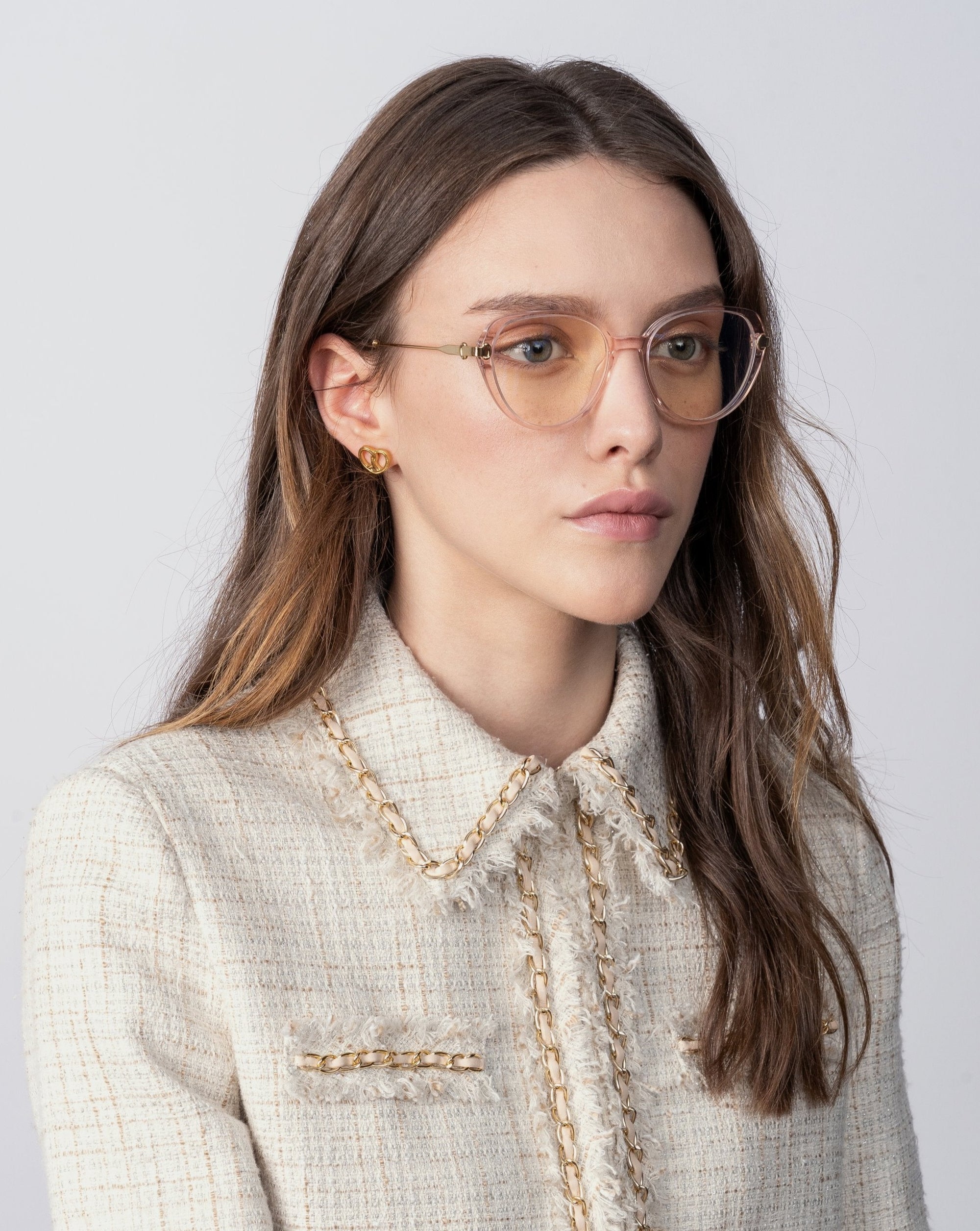 A woman with long, light brown hair is wearing a cream-colored tweed jacket with 18-karat gold-plated accents and a pair of For Art&#39;s Sake® Waterhouse clear-framed eyeglasses. She appears to be looking slightly to her left with a neutral expression against a plain white background.