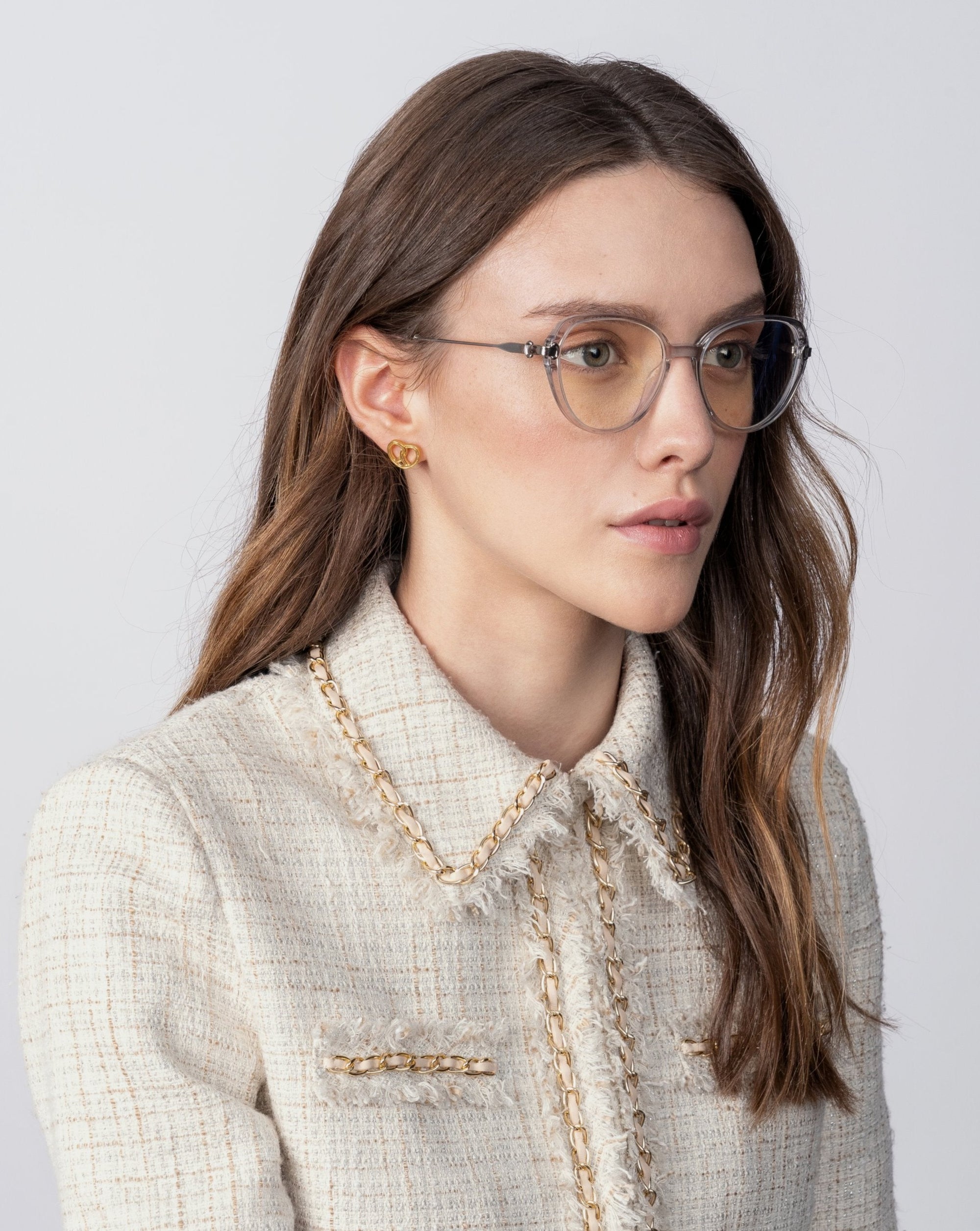 A woman with long, wavy brown hair is wearing rectangular eyeglasses called Waterhouse by For Art&#39;s Sake® with prescription lenses and gold earrings. She is dressed in a cream-colored, textured jacket with decorative trim. She gazes slightly to the right against a plain, light background.