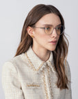 A woman with long, wavy brown hair is wearing rectangular eyeglasses called Waterhouse by For Art's Sake® with prescription lenses and gold earrings. She is dressed in a cream-colored, textured jacket with decorative trim. She gazes slightly to the right against a plain, light background.