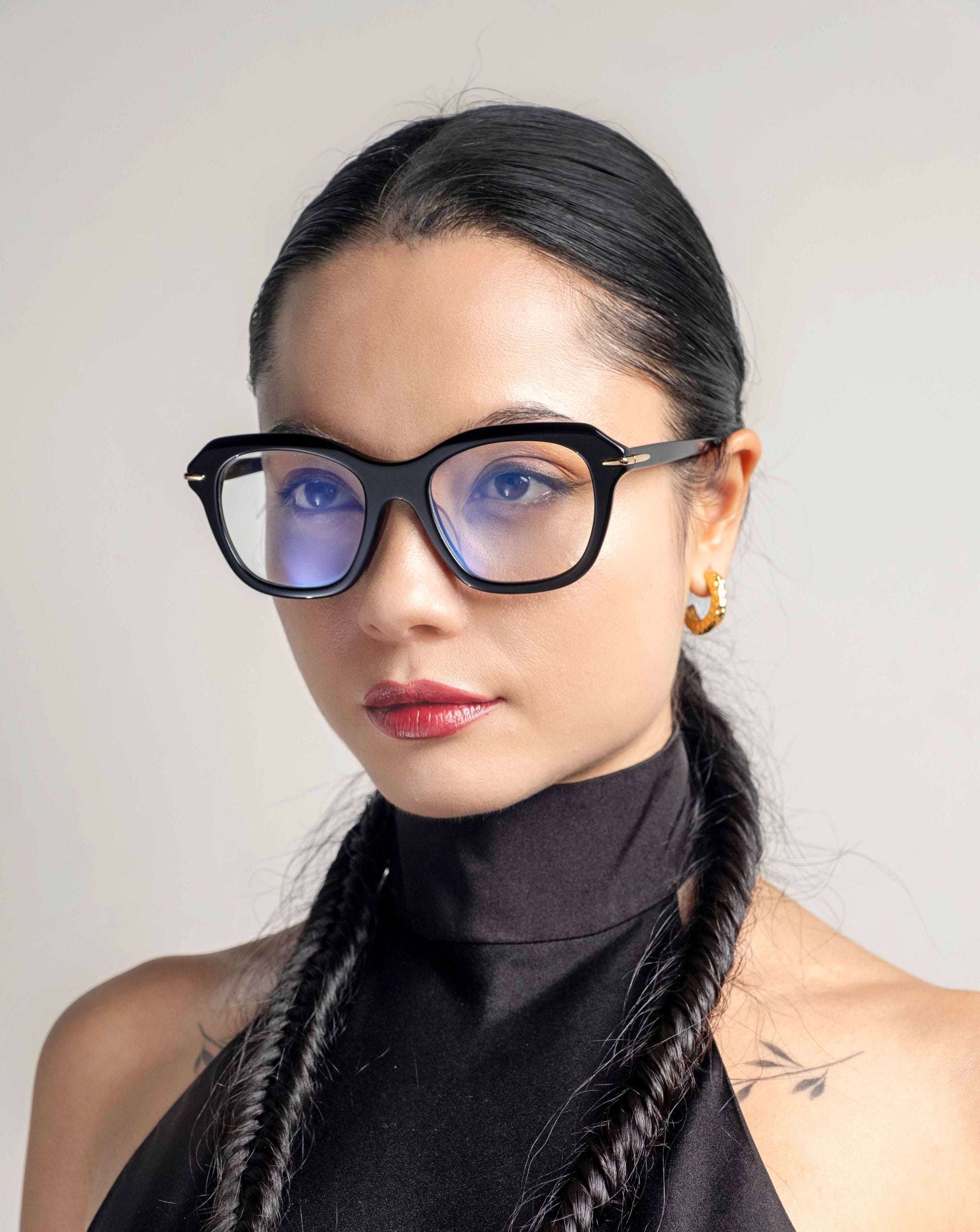 A person with long, dark hair styled in twin braids wears black-framed glasses with blue lenses. They are dressed in a black high-neck outfit and have small 18-karat gold-plated Helene hoop earrings by For Art's Sake®. The background is neutral, focusing attention on the person.