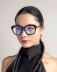 A person with long, dark hair styled in twin braids wears black-framed glasses with blue lenses. They are dressed in a black high-neck outfit and have small 18-karat gold-plated Helene hoop earrings by For Art's Sake®. The background is neutral, focusing attention on the person.