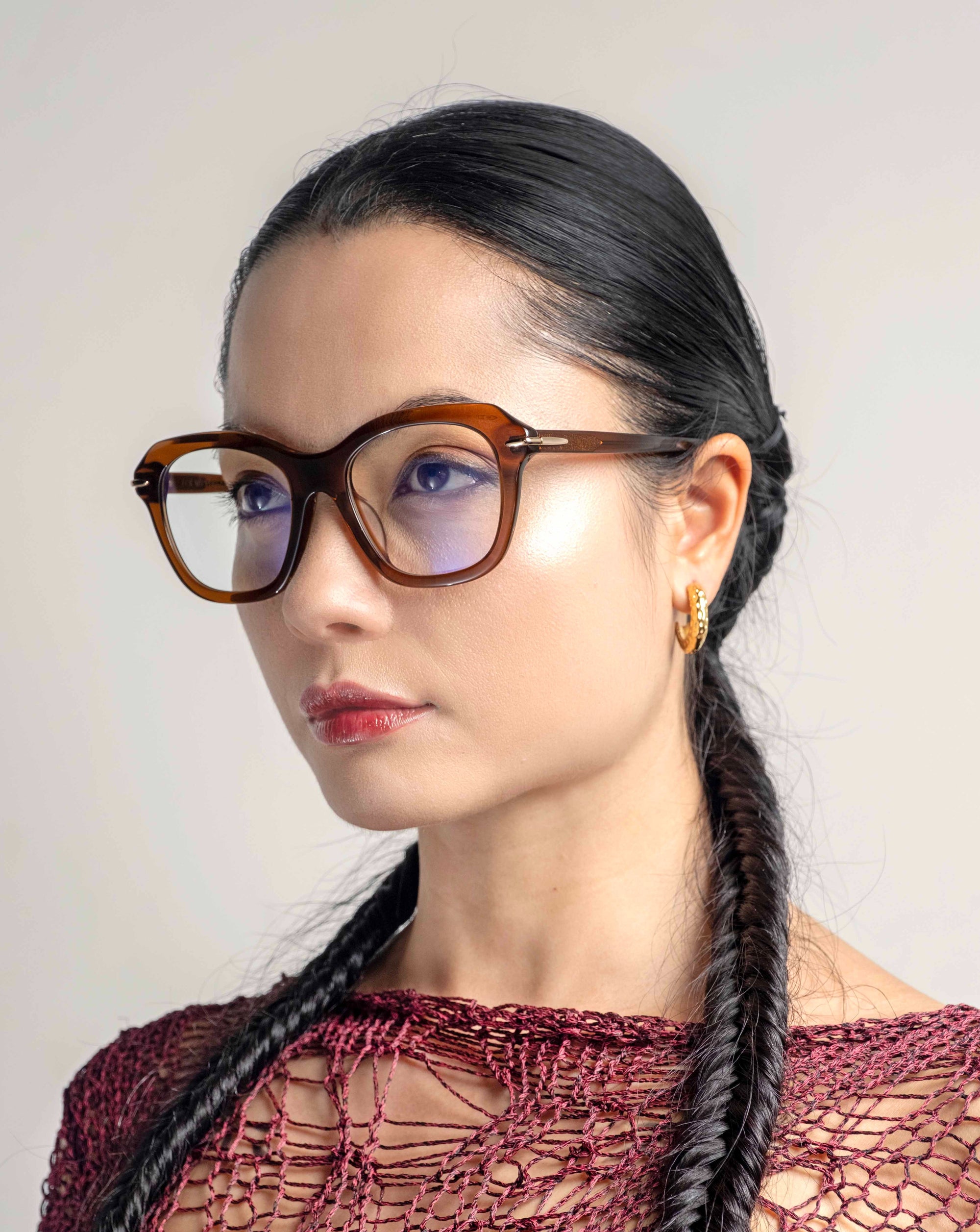 A woman with long, dark hair styled in two braids wears large, brown-framed Helene glasses with a cat-eye silhouette from For Art&#39;s Sake® and a gold hoop earring. She has a neutral expression and is dressed in a red, textured garment, with a light gray background behind her.