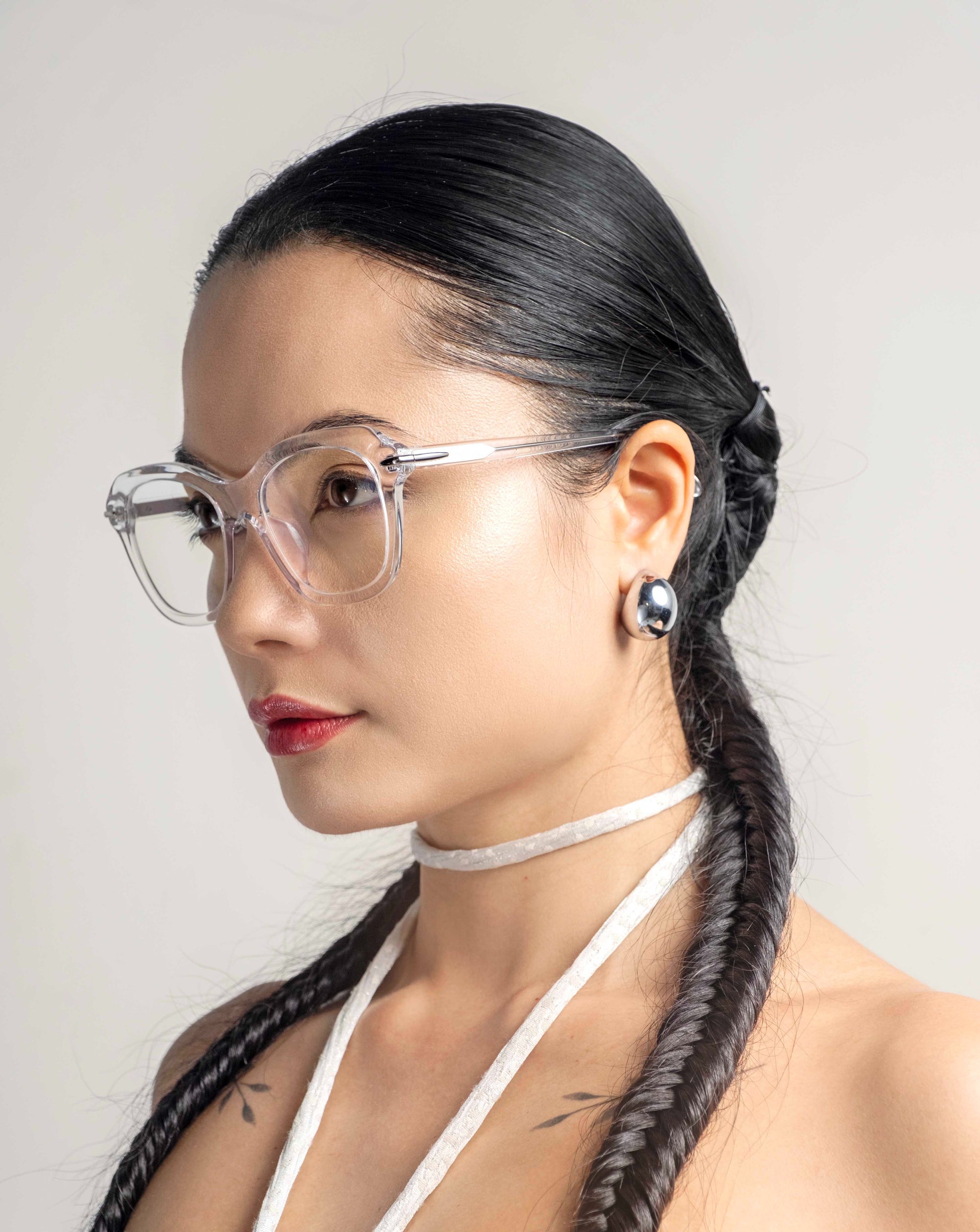 A person with long, dark braided hair, wearing clear-framed, cat-eye silhouette glasses and a white halter strap, is pictured in a profile view against a plain background. They have a calm expression with shiny Helene earrings by For Art&#39;s Sake® visible.