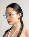 A person with long, dark braided hair, wearing clear-framed, cat-eye silhouette glasses and a white halter strap, is pictured in a profile view against a plain background. They have a calm expression with shiny Helene earrings by For Art's Sake® visible.