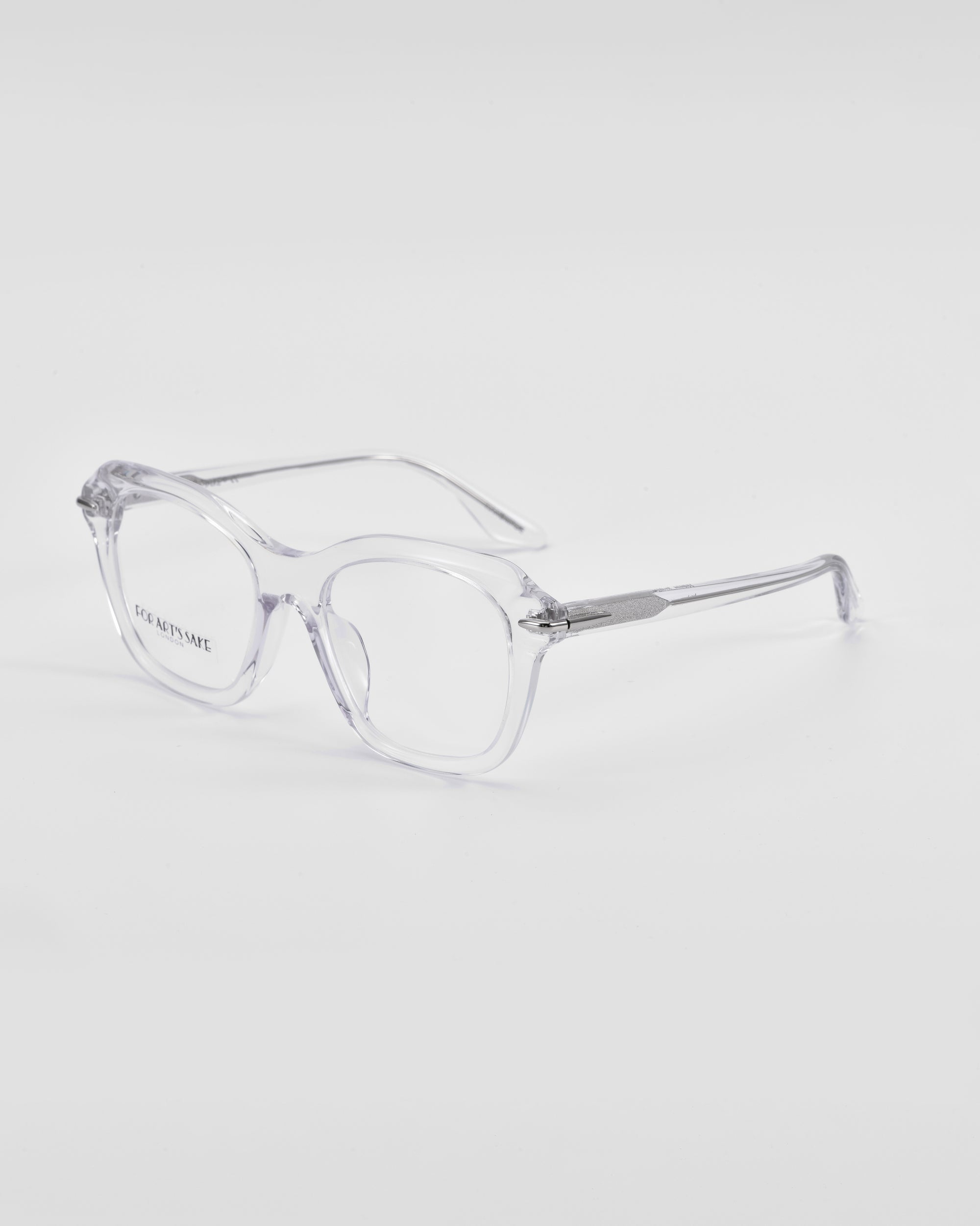 A pair of clear, transparent Helene by For Art&#39;s Sake® eyeglasses with square frames is placed on a white surface. The lenses are large with a slight retro style, and the temples feature 18-karat gold plating near the hinges. The glasses have a simple and minimalist design perfect for those seeking both elegance and functionality.
