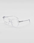 A pair of clear, transparent Helene by For Art's Sake® eyeglasses with square frames is placed on a white surface. The lenses are large with a slight retro style, and the temples feature 18-karat gold plating near the hinges. The glasses have a simple and minimalist design perfect for those seeking both elegance and functionality.