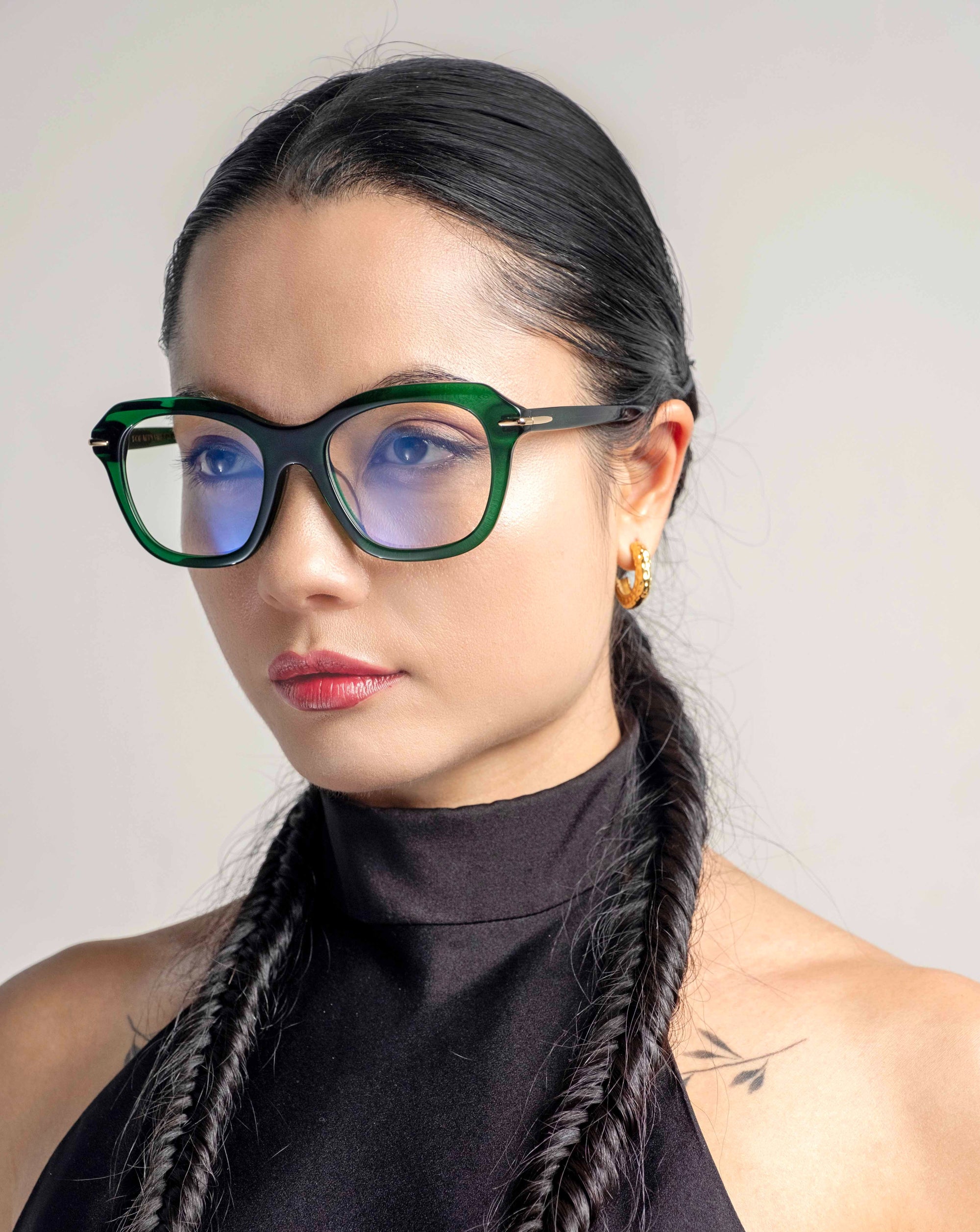 A person with long, dark, braided hair wears large, cat-eye silhouette glasses named Helene from For Art&#39;s Sake® with blue-tinted lenses. They are dressed in a black, high-neck top and have small hoop earrings. The background is neutral.