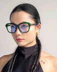 A person with long, dark, braided hair wears large, cat-eye silhouette glasses named Helene from For Art's Sake® with blue-tinted lenses. They are dressed in a black, high-neck top and have small hoop earrings. The background is neutral.