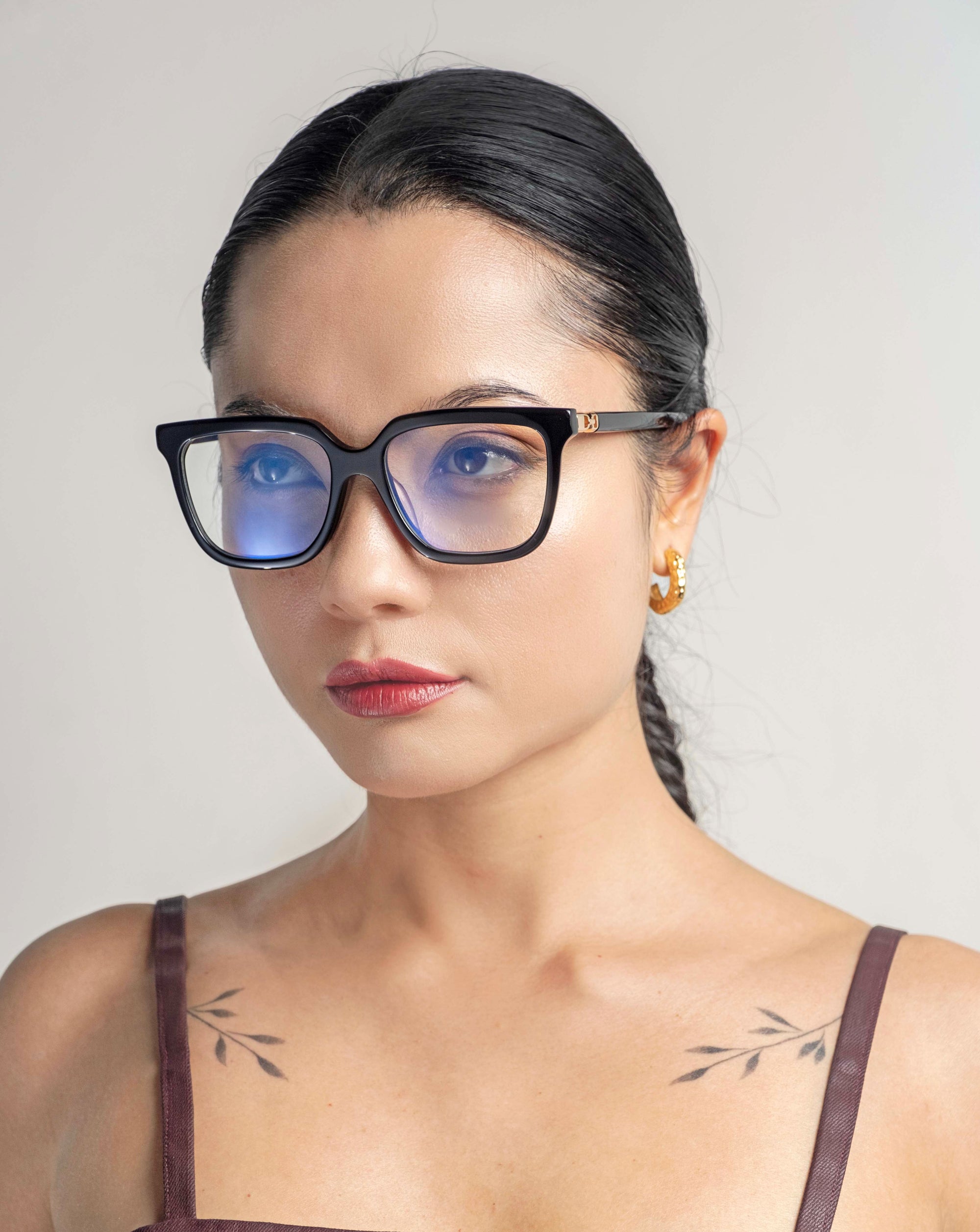 A person with long black hair, styled in a braid, is wearing Nina glasses from For Art's Sake® and 18-karat gold hoop earrings. They have subtle tattoos on their shoulders, visible due to the sleeveless top they are wearing. The background is light and plain.