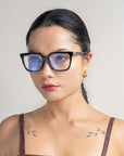 A person with long black hair, styled in a braid, is wearing Nina glasses from For Art's Sake® and 18-karat gold hoop earrings. They have subtle tattoos on their shoulders, visible due to the sleeveless top they are wearing. The background is light and plain.