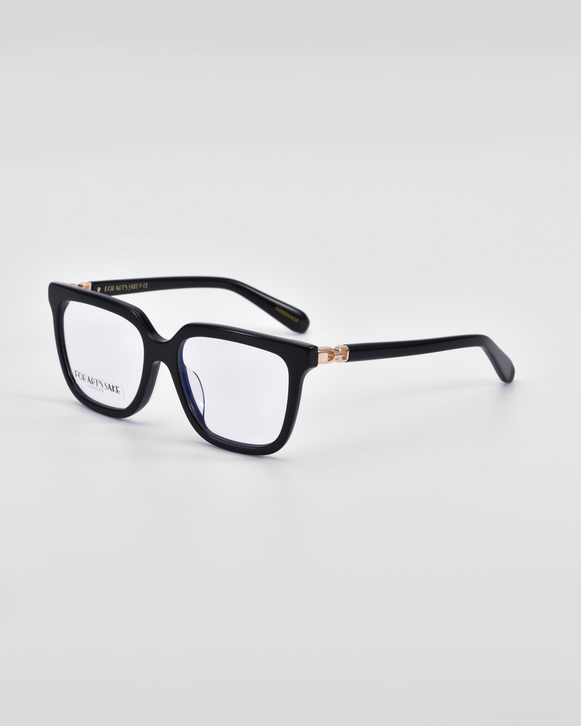 A pair of black rectangular eyeglasses with thick frames is shown on a plain white background. The glasses feature clear lenses and a small 18-karat gold hinge detail on the temples, combining luxury with a classic square silhouette. These are the Nina by For Art&#39;s Sake®.