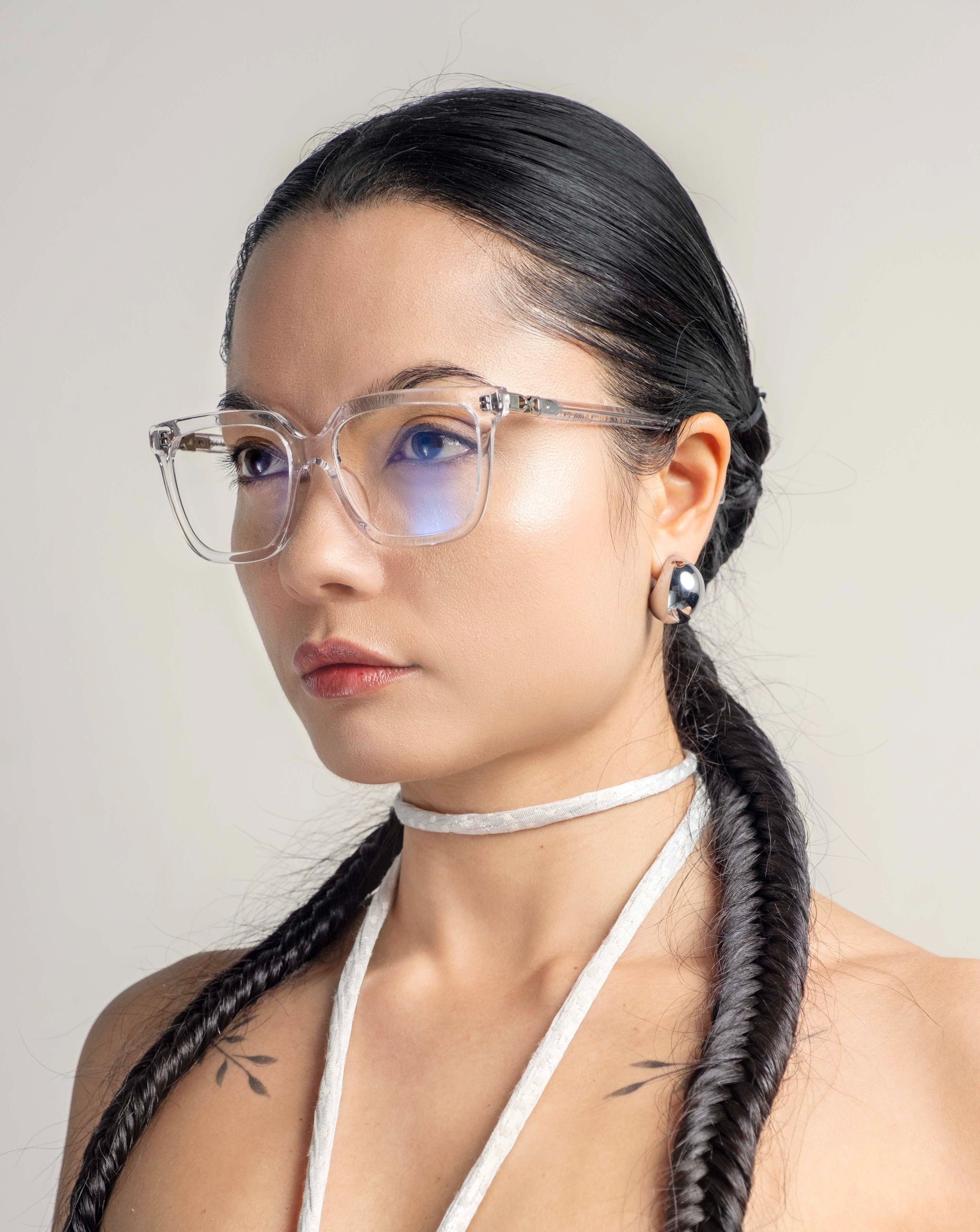 A person with long, dark braided hair is wearing For Art's Sake® Nina glasses and a white choker. They have a serious expression and are dressed in a light-colored top. The classic square silhouette of the glasses adds to their poised look, set against a plain, neutral background.