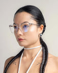 A person with long, dark braided hair is wearing For Art's Sake® Nina glasses and a white choker. They have a serious expression and are dressed in a light-colored top. The classic square silhouette of the glasses adds to their poised look, set against a plain, neutral background.