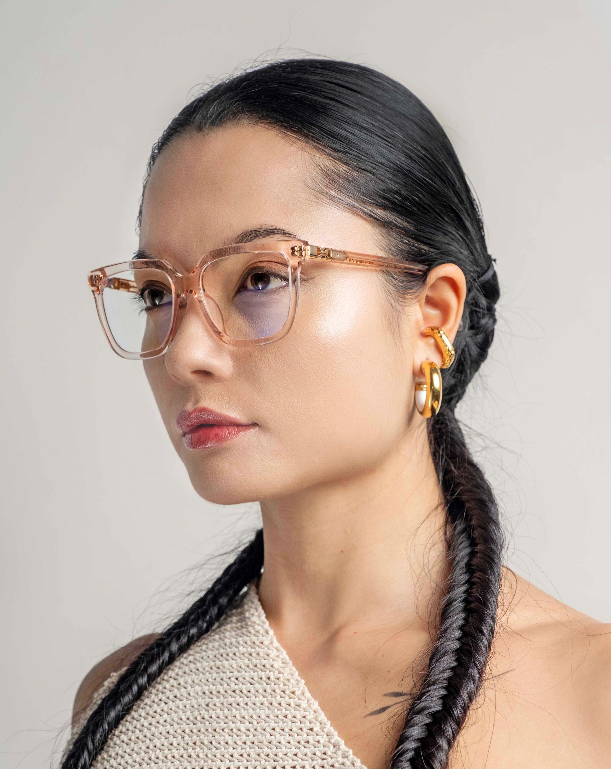 A woman with long, braided hair is seen wearing For Art&#39;s Sake® Nina transparent eyeglasses in a classic square silhouette and 18-karat gold hoop earrings. She is dressed in a textured off-white one-shoulder top and is looking slightly to her right against a plain background. The overall mood is stylish and poised.