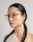 A woman with long, braided hair is seen wearing For Art's Sake® Nina transparent eyeglasses in a classic square silhouette and 18-karat gold hoop earrings. She is dressed in a textured off-white one-shoulder top and is looking slightly to her right against a plain background. The overall mood is stylish and poised.