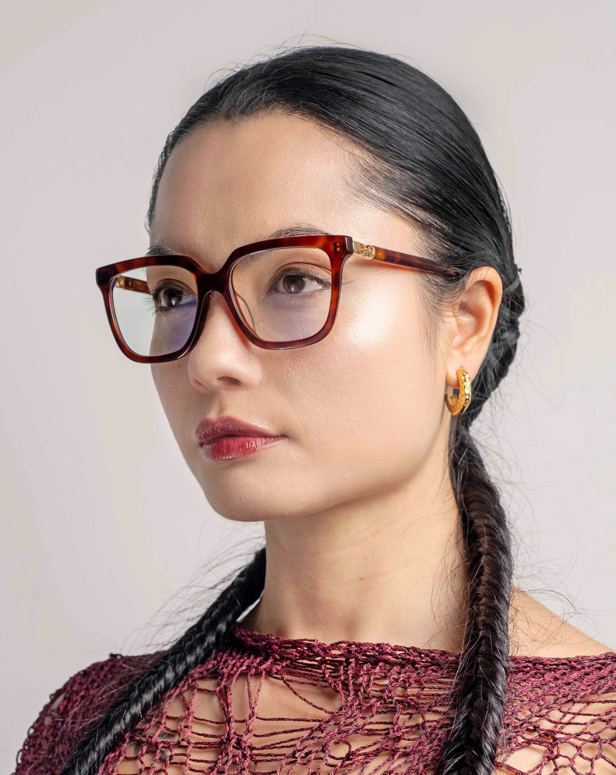 A woman with long black hair styled in two braids is wearing large, tortoiseshell For Art&#39;s Sake® Nina glasses in a classic square silhouette and 18-karat gold hoop earrings. She is dressed in a red, intricately woven top and is posed against a neutral background, looking towards the left.