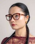 A woman with long black hair styled in two braids is wearing large, tortoiseshell For Art's Sake® Nina glasses in a classic square silhouette and 18-karat gold hoop earrings. She is dressed in a red, intricately woven top and is posed against a neutral background, looking towards the left.