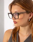 A person with long, light brown hair wears oversized, square-framed Abbey glasses from For Art's Sake® and drop earrings. They are looking to their left and are dressed in a sleeveless, dark-colored top adorned with a subtle link-chain pattern. The background is plain and light.