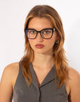 A person with long, light brown hair is wearing black-rimmed glasses and a gray sleeveless top. The Abbey by For Art's Sake® frames their neutral expression perfectly, set against a plain white background.