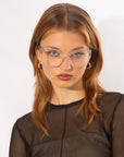 A person with light brown hair and clear cat-eye Abbey opticals by For Art's Sake® is pictured against a plain white background. They are wearing a sheer black top and gold hoop earrings, looking directly into the camera with a neutral expression.