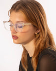 A person with long, light brown hair is shown in profile wearing large, clear-framed glasses. They have gold hoop earrings and are dressed in a dark top. The background is plain white. Their stylish eyewear hints at an Abbey by For Art's Sake® opticals prescription service designed for both fashion and function.
