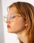 A person with light brown hair is shown in profile wearing oversized, transparent eyeglasses with 18-karat gold plated details. They are also wearing Abbey by For Art's Sake® and have a neutral expression. The background is plain white.