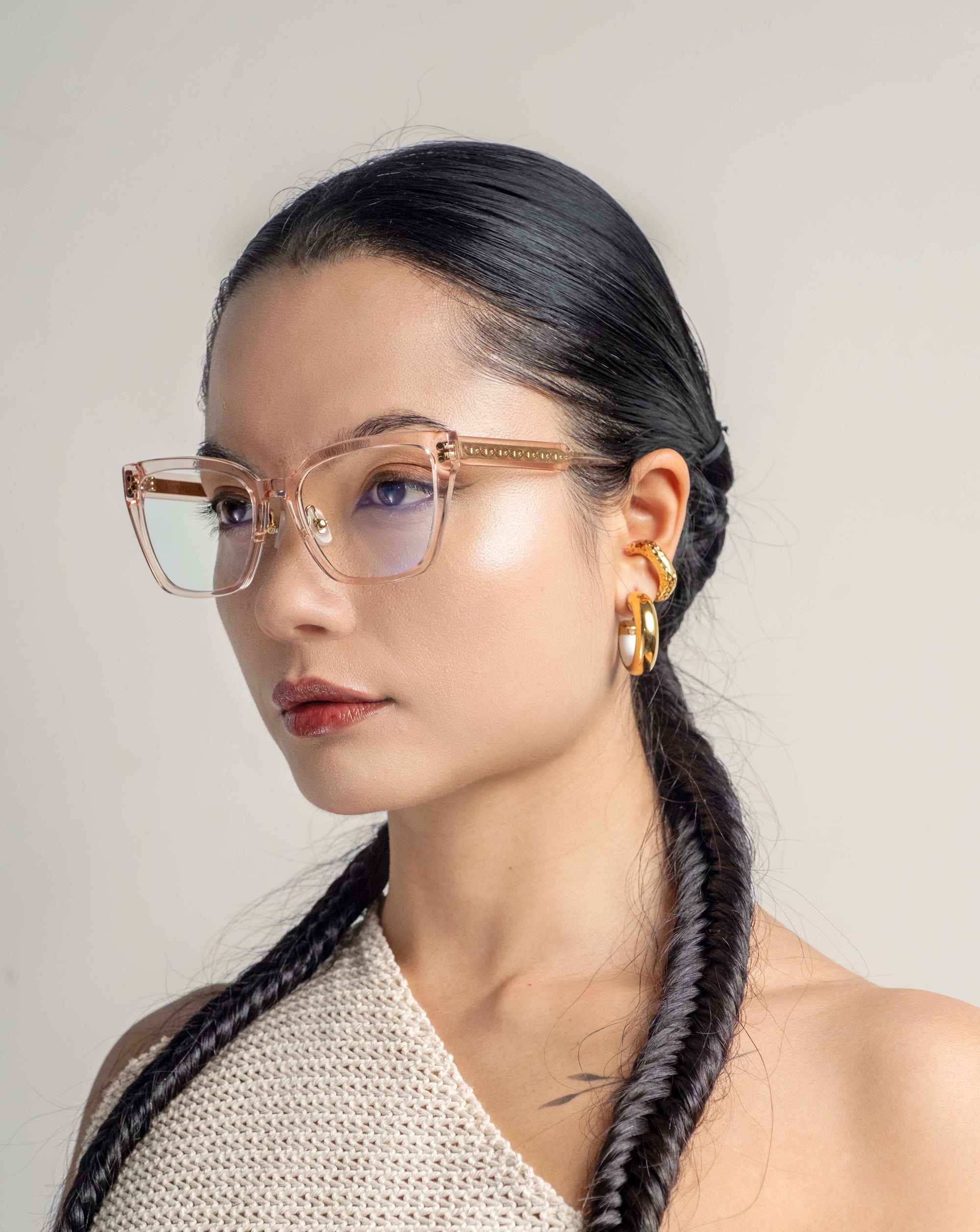 A person with long, dark braided hair is wearing square, translucent **Abbey** eyeglasses from **For Art&#39;s Sake®** and bold, gold hoop earrings. They are dressed in a textured, off-white, sleeveless garment with a subtle link-chain pattern against a plain background, giving a minimalist and sophisticated appearance.