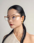 A person with long, dark braided hair is wearing square, translucent **Abbey** eyeglasses from **For Art's Sake®** and bold, gold hoop earrings. They are dressed in a textured, off-white, sleeveless garment with a subtle link-chain pattern against a plain background, giving a minimalist and sophisticated appearance.