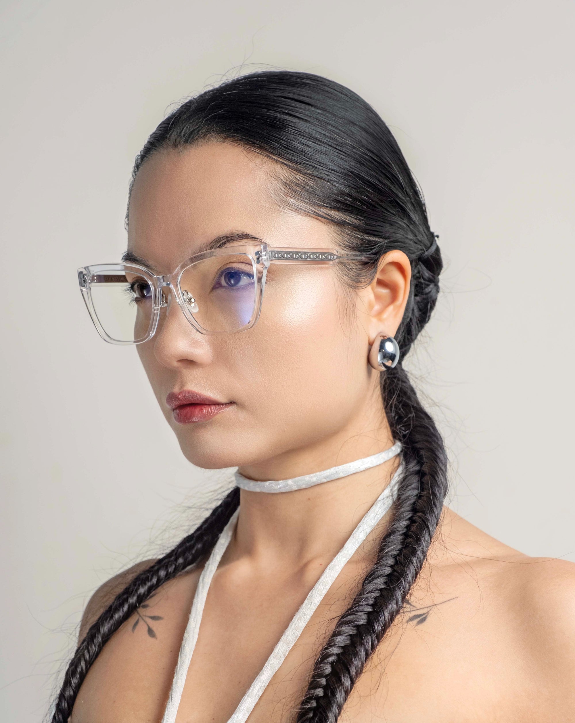 A woman with long, dark hair styled in twin braids and wearing Abbey clear-framed glasses by For Art&#39;s Sake® looks to the side. She has small tattoos on her shoulders, wears large, circular 18-karat gold plated earrings, and a white choker. The background is plain and light-colored.