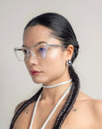 A woman with long, dark hair styled in twin braids and wearing Abbey clear-framed glasses by For Art's Sake® looks to the side. She has small tattoos on her shoulders, wears large, circular 18-karat gold plated earrings, and a white choker. The background is plain and light-colored.