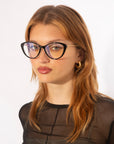 A woman with long brown hair is wearing large, black-rimmed For Art's Sake® Perla II glasses with jade-stone nose pads and gold hoop earrings. She is dressed in a black mesh top and is looking slightly to the side against a plain white background.