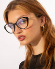 A person with long, light brown hair is wearing large, black cat-eye glasses featuring jade-stone nose pads and small gold hoop earrings. They are looking slightly to the side with a neutral expression on their face. The background is plain white. The glasses are Perla II by For Art's Sake®.