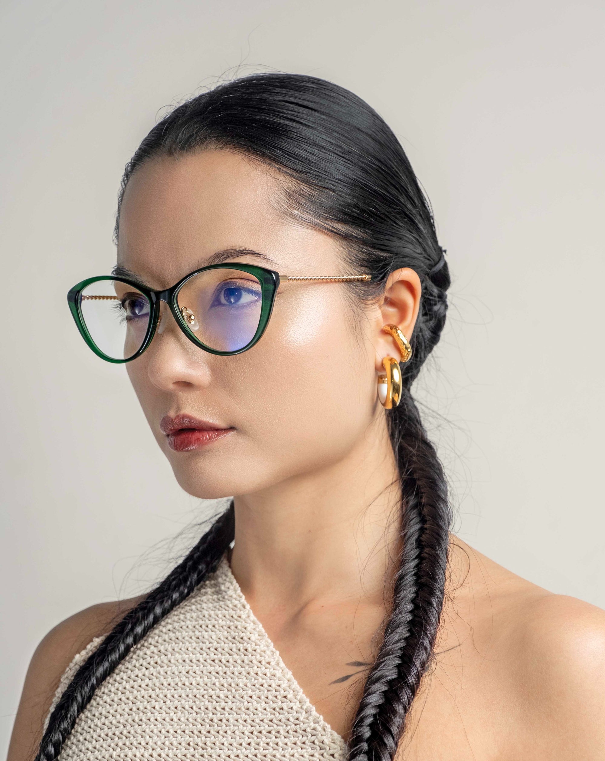 A woman with long, dark hair styled in a braided ponytail wears black cat-eye Perla II glasses by For Art&#39;s Sake® featuring blue-tinted lenses and 18-karat gold plating. She has gold hoop earrings and is dressed in a knitted beige top while standing against a plain light-colored background.